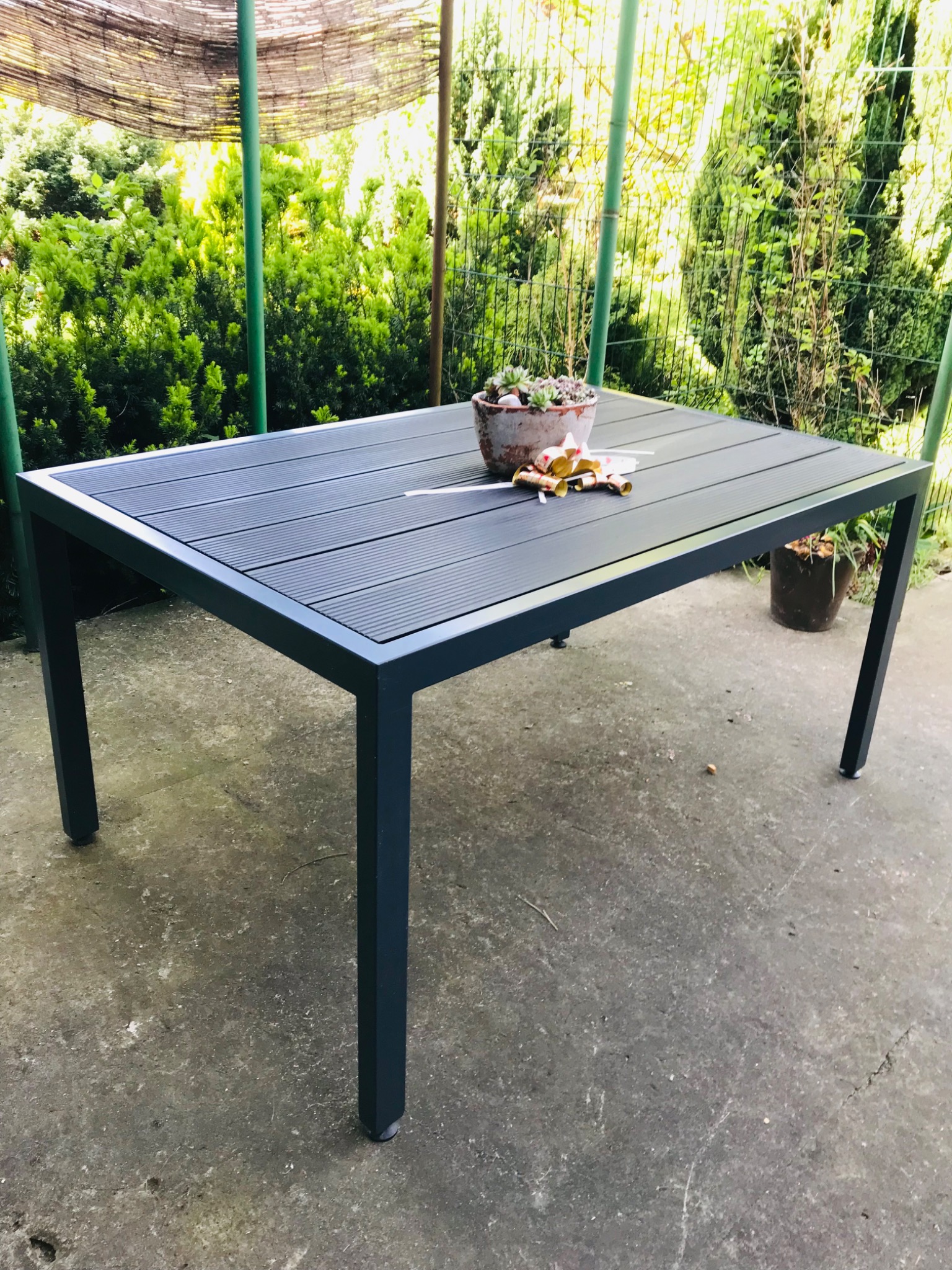 Stół ogrodowy z metalową ramą i blatem z desek kompozytowych, stojący na betonowym podłożu w Kłobucku, z doniczką z sukulentami na blacie i zielenią w tle.