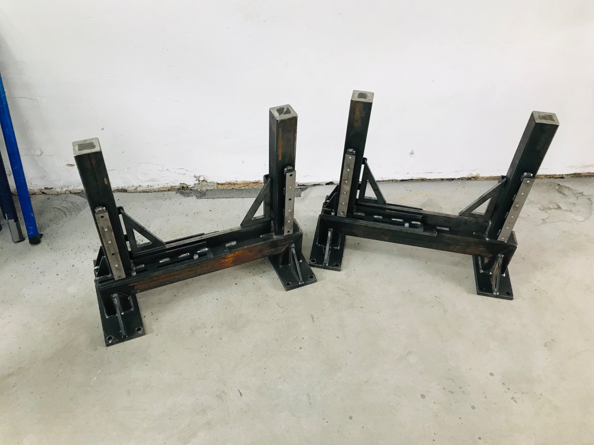 Dwa metalowe stojaki spawalnicze z kwadratowych profili, ustawione na betonowej posadzce przy białej ścianie, widoczne ślady spawania i niedoskonałości wykończenia.