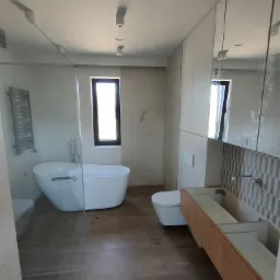 Nowoczesna łazienka z wolnostojącą wanną, przeszklonym prysznicem, podwieszaną toaletą i szafką z umywalką na tle geometrycznej mozaiki.