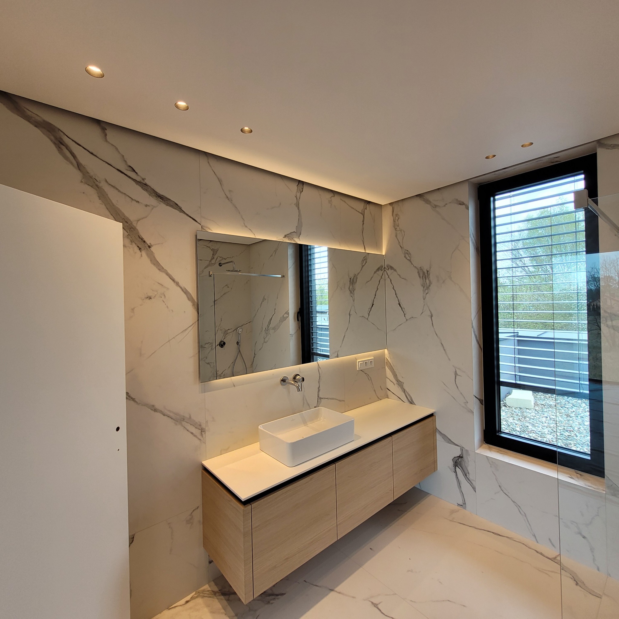 Nowoczesna łazienka z marmurowymi płytkami, podświetlanym lustrem i drewnianą szafką pod umywalką, widok na zewnątrz przez okno z żaluzjami.