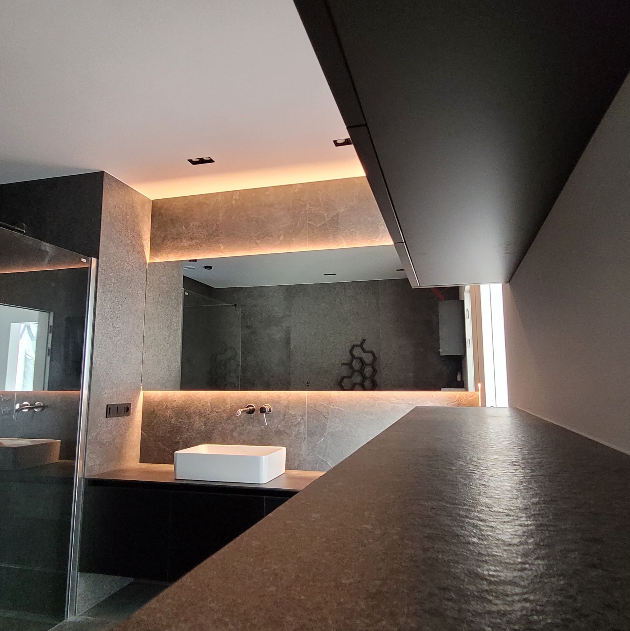 Nowoczesna łazienka w odcieniach szarości z podświetlanym lustrem, białą umywalką nablatową i czarnymi szafkami, widok z perspektywy blatu z ciemnego kamienia.