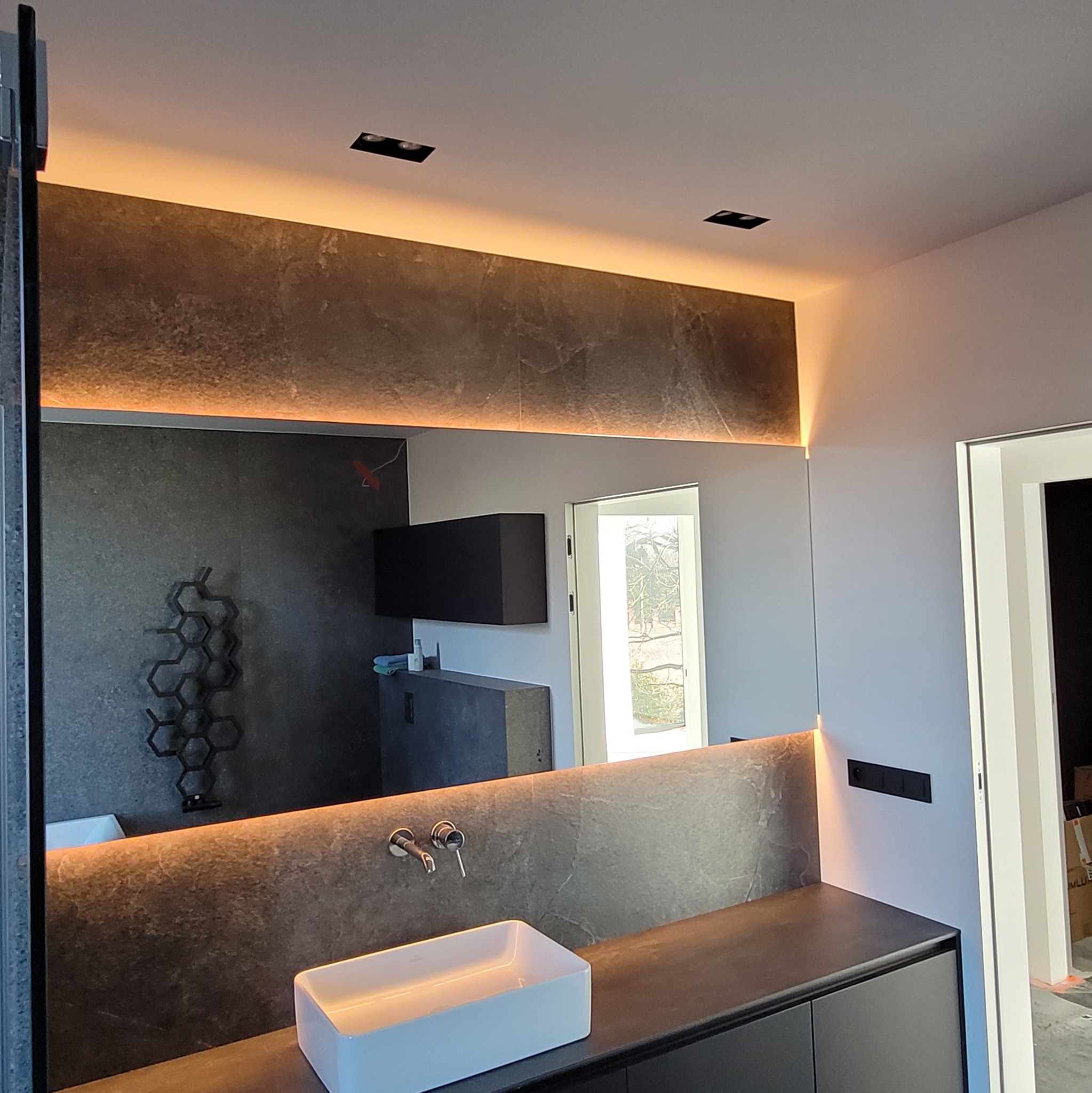 Nowoczesna łazienka z podświetlanym lustrem, grafitowa ściana z dekoracyjnym grzejnikiem, minimalistyczna umywalka nablatowa i szafka pod umywalką w kolorze czarnym, widoczne elementy wykończeniowe.