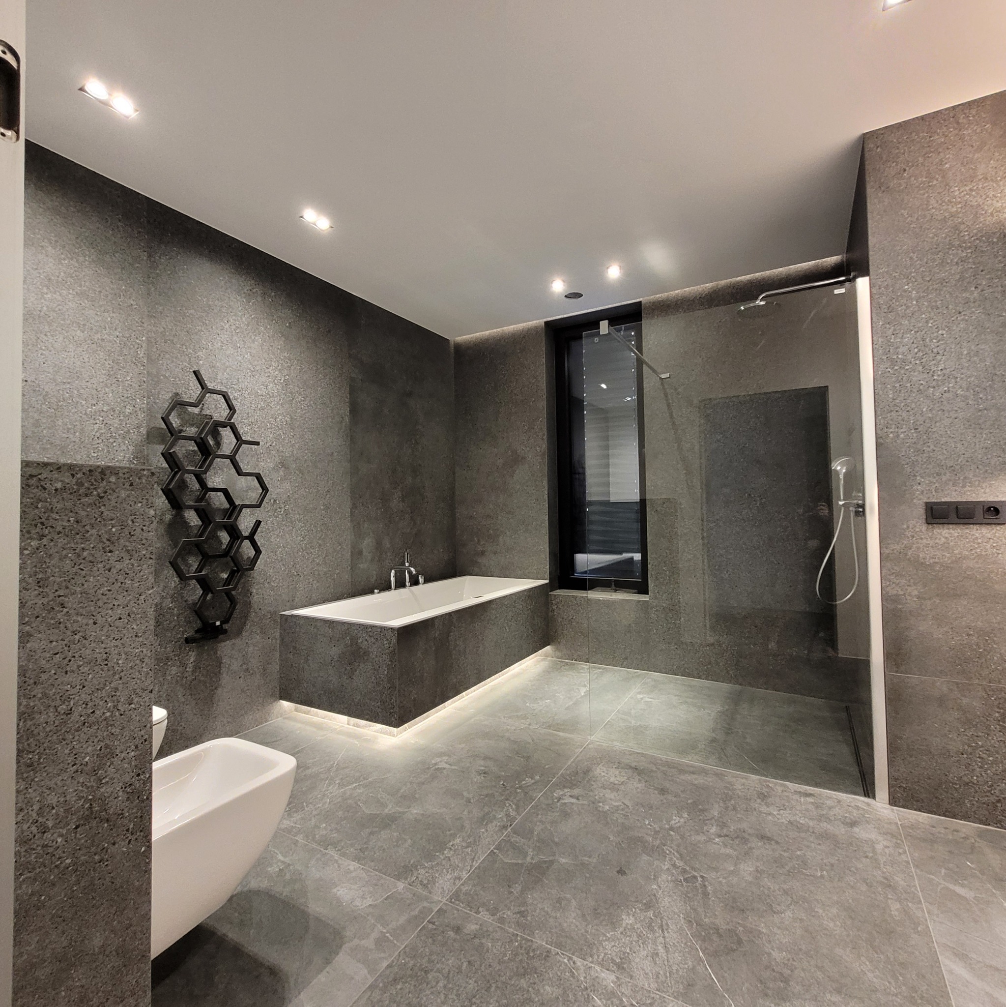 Nowoczesna łazienka w odcieniach szarości z podświetloną wanną, grzejnikiem w kształcie plastra miodu i przeszkloną kabiną prysznicową.