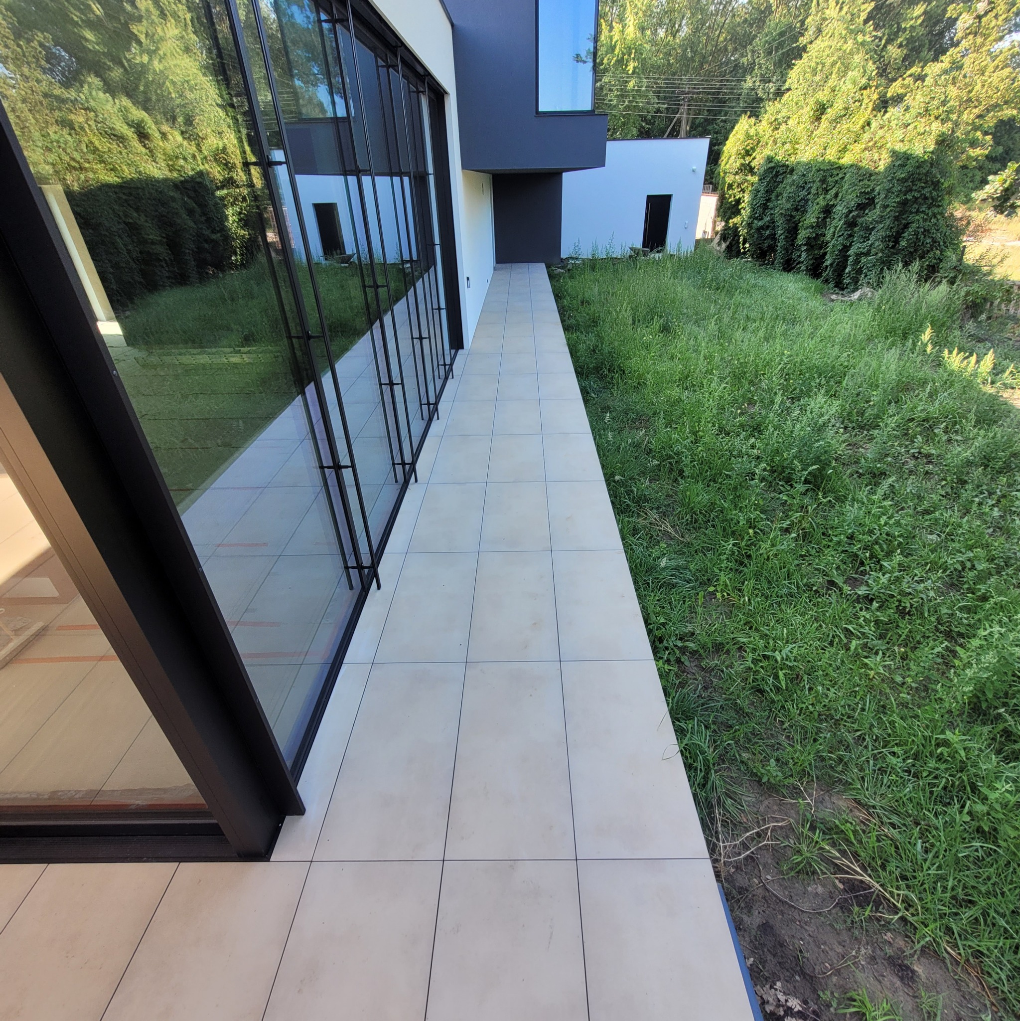 Jasne płytki podłogowe na zewnątrz nowoczesnego budynku w Pabianicach, widok na bujną trawę i odbicie zieleni w dużych oknach.