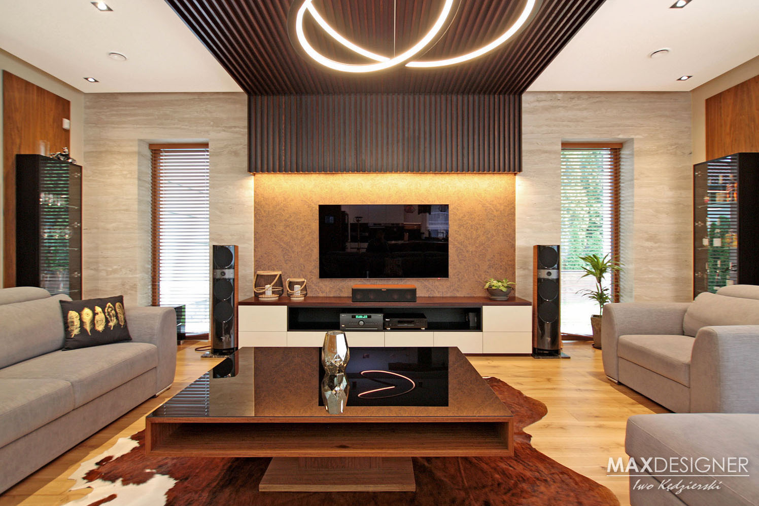 Salon z drewnianą podłogą, szarymi sofami z poduszkami, stolikiem kawowym z ciemnego drewna i szklanym blatem, telewizorem na ścianie, głośnikami po bokach, oraz dekoracyjnym oświetleniem sufitowym...