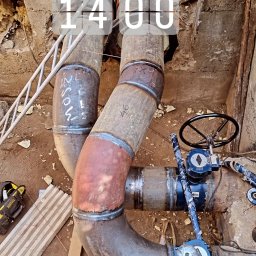 Łukasz Blitek - Zespawane rury w wykopie, z widocznymi zaworami i elementami mocującymi, częściowo pokryte izolacją, z napisem '14:00' nałożonym na zdjęcie.