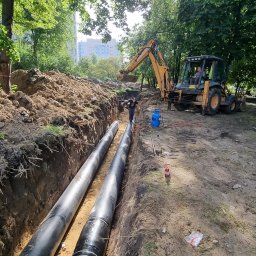 Łukasz Blitek - Dwie czarne rury ułożone w wykopie ziemnym, pracownik stoi na jednej z nich, w tle żółta koparka i zieleń drzew.