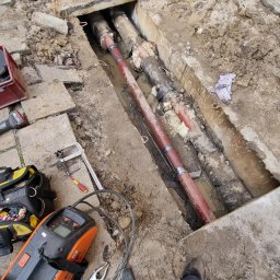 Łukasz Blitek - Odkryte rury w wykopie na posesji w Koniuszy, widoczne narzędzia spawalnicze i fragment stroju roboczego. Rury częściowo zabezpieczone czerwoną powłoką antykorozyjną.