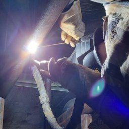 Łukasz Blitek - Prace spawalnicze na wysokości: spawanie rury stalowej w trudno dostępnym miejscu, widoczna iskra i ochronna maska na twarzy pracownika, rękawice robocze i ubranie robocze.