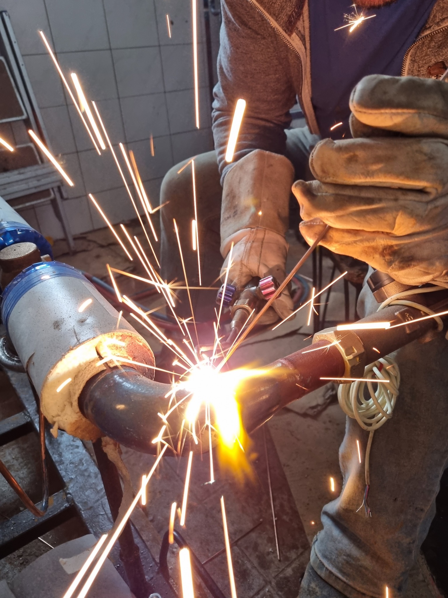 Spawanie rury z gwintem w warsztacie, widoczne iskry, brudne rękawice robocze i fragment instalacji.