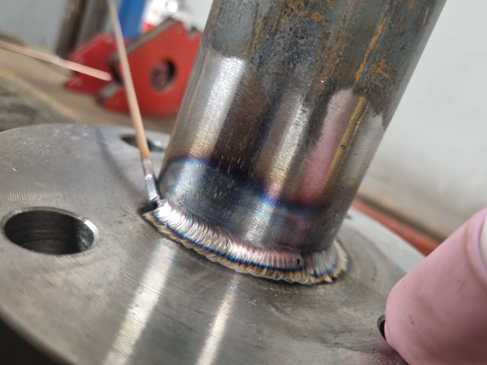 Detal spoiny TIG łączącej rurę ze stalową flanszą, widoczne tęczowe zabarwienia na spoinie, elektroda wolframowa oraz uchwyt spawalniczy.