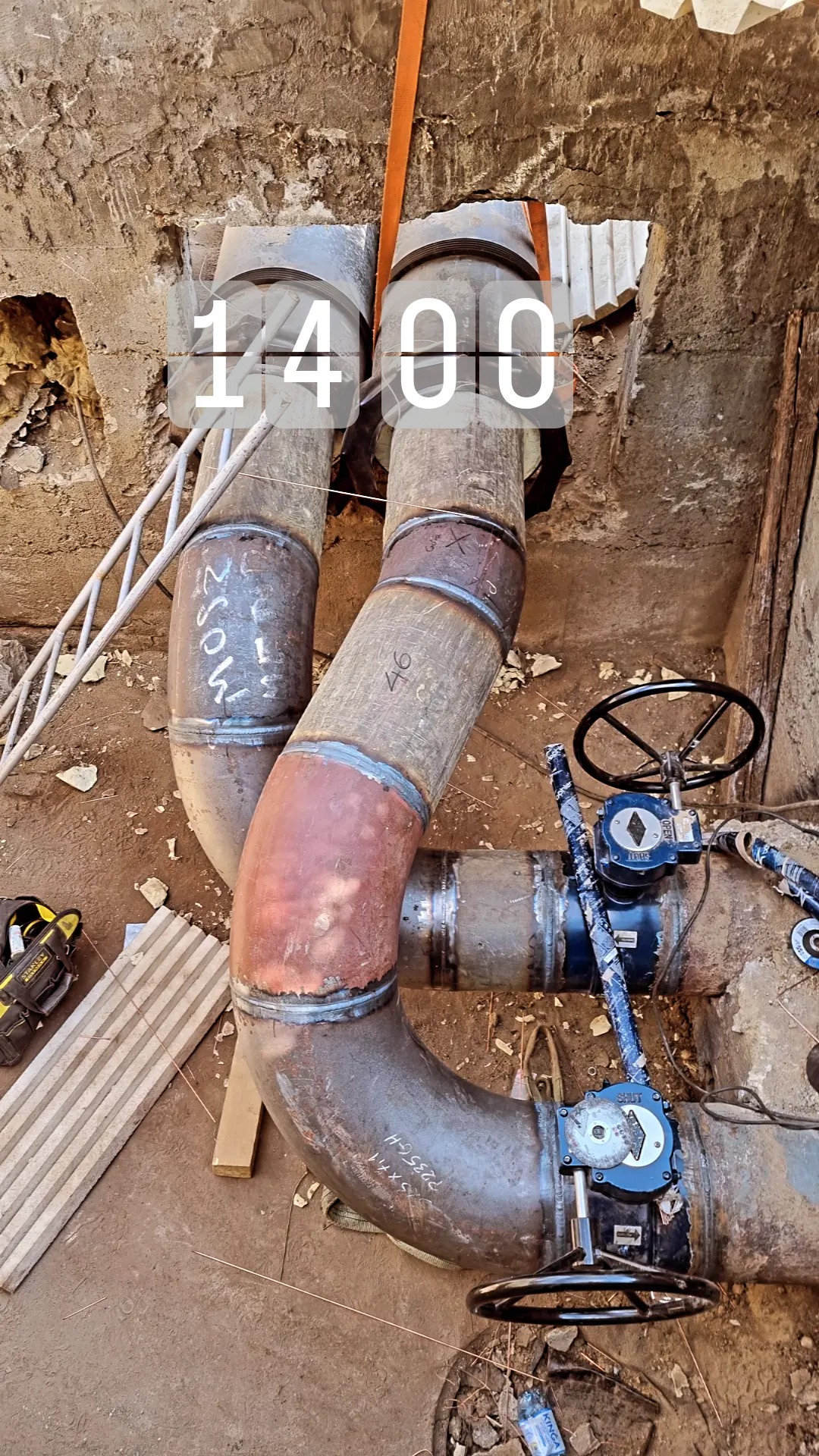 Zespawane rury w wykopie, z widocznymi zaworami i elementami mocującymi, częściowo pokryte izolacją, z napisem '14:00' nałożonym na zdjęcie.