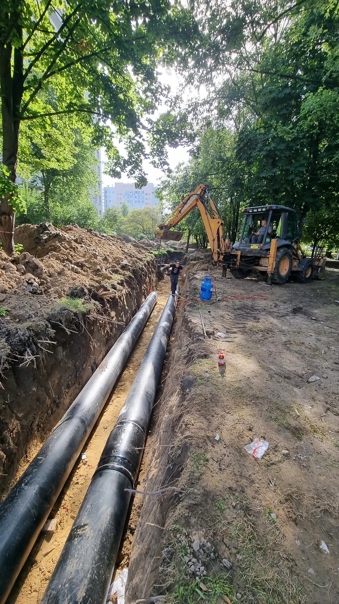 Dwie czarne rury ułożone w wykopie ziemnym, pracownik stoi na jednej z nich, w tle żółta koparka i zieleń drzew.