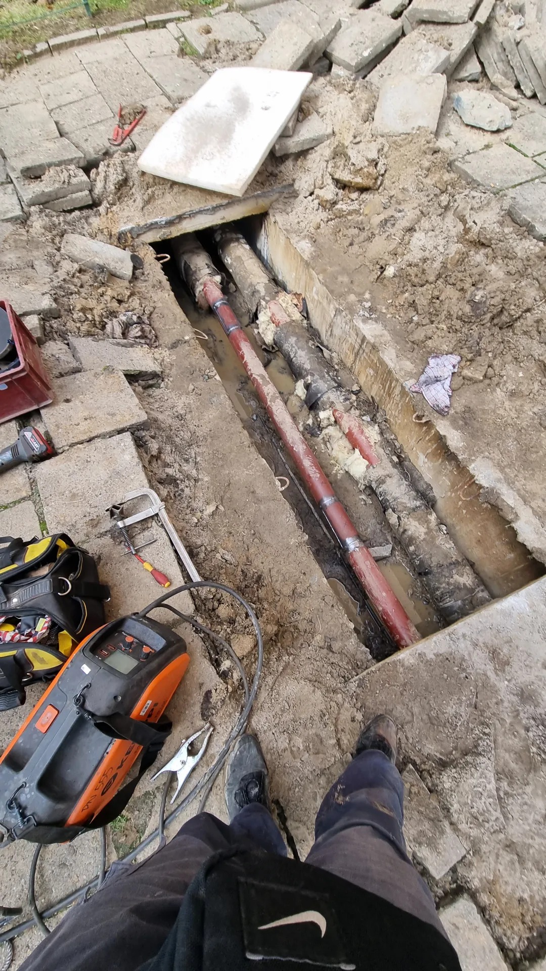 Odkryte rury w wykopie na posesji w Koniuszy, widoczne narzędzia spawalnicze i fragment stroju roboczego. Rury częściowo zabezpieczone czerwoną powłoką antykorozyjną.