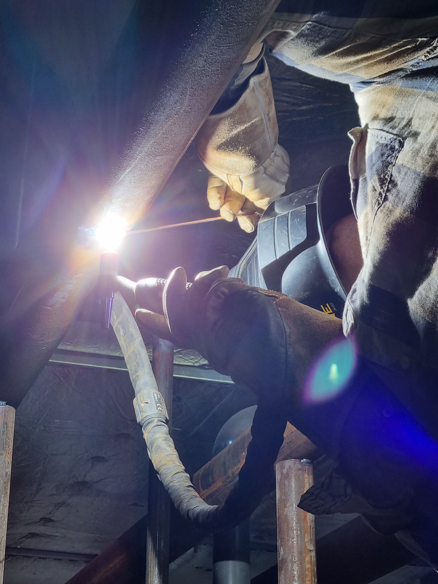 Prace spawalnicze na wysokości: spawanie rury stalowej w trudno dostępnym miejscu, widoczna iskra i ochronna maska na twarzy pracownika, rękawice robocze i ubranie robocze.