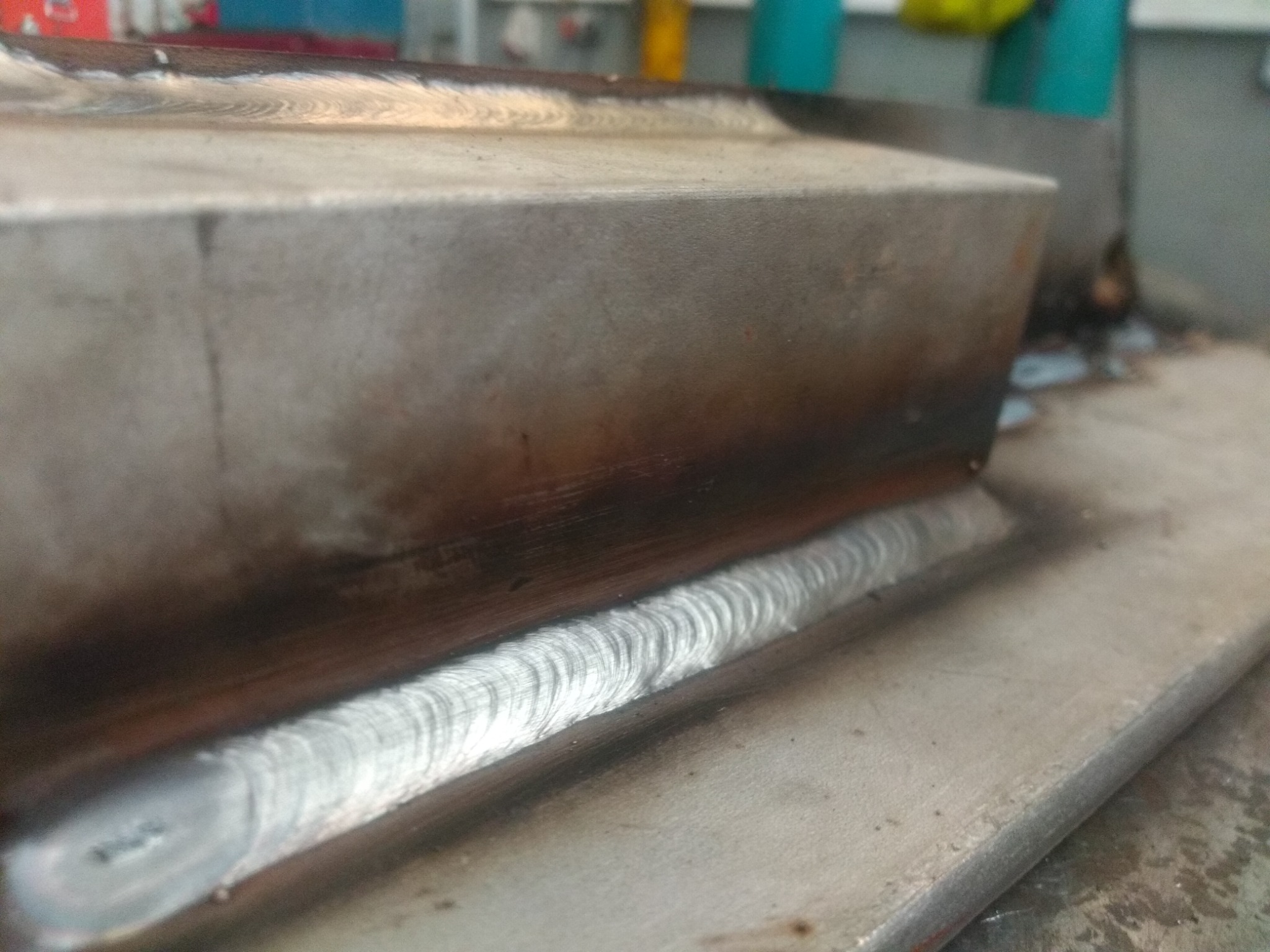 Metalowy element po spawaniu z widocznym świeżym spawem o charakterystycznym wzorze, leżący na metalowej powierzchni roboczej.