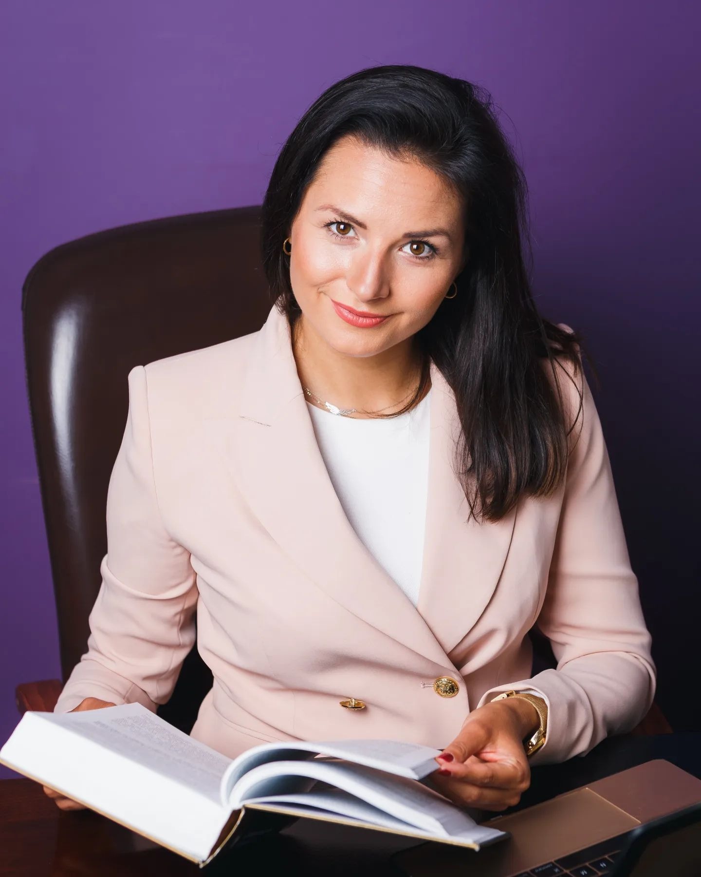 Uśmiechnięta prawniczka w różowym żakiecie siedzi przy biurku, czytając otwartą książkę, z laptopem obok na tle fioletowej ściany.
