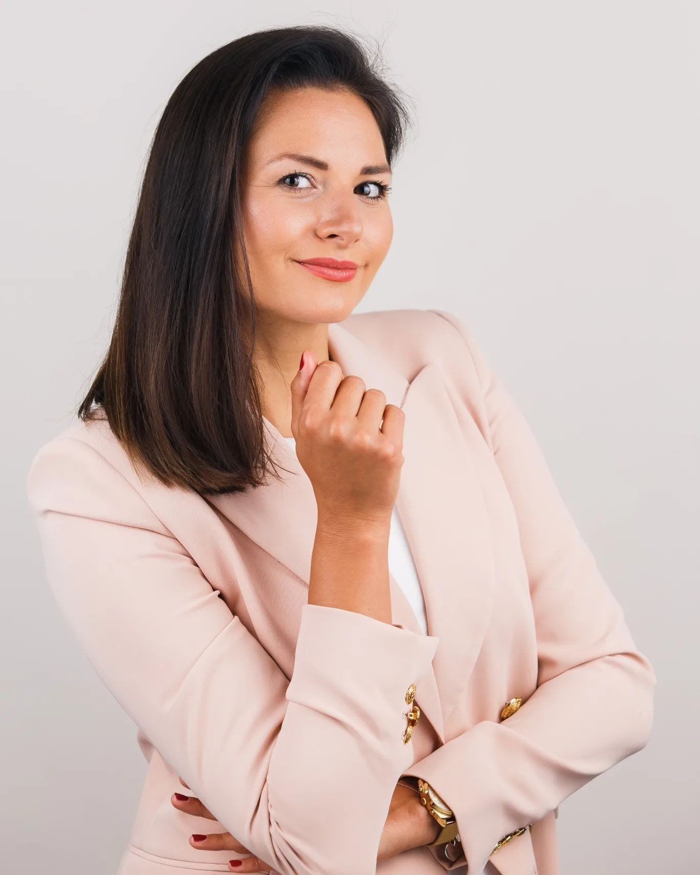 Portret uśmiechniętej prawniczki z Warszawy w pudrowym żakiecie, z ręką pod brodą i złotym zegarkiem, pozującej na jasnym tle.