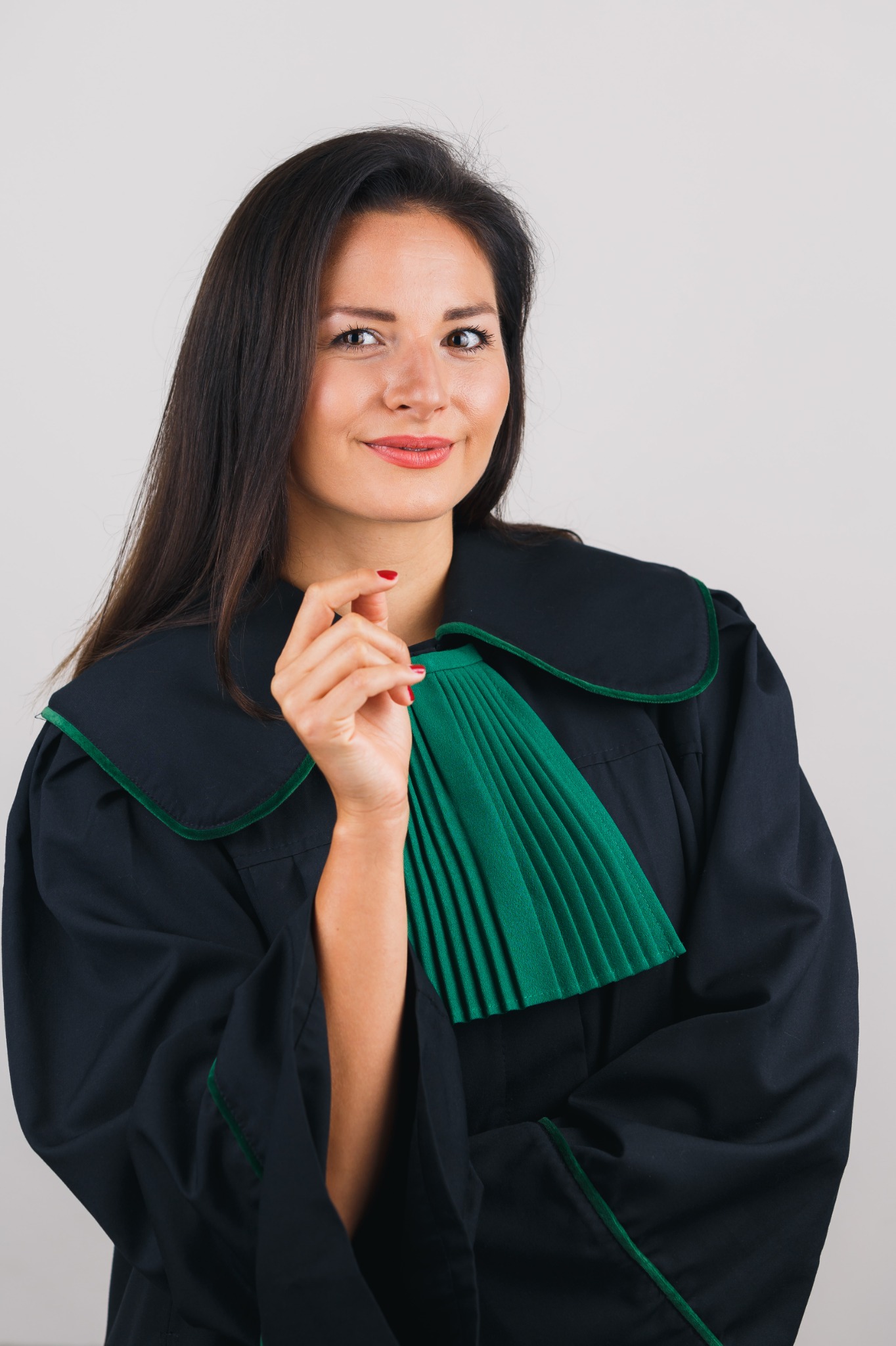 Uśmiechnięta brunetka w todze adwokackiej z zielonym żabotem, trzymająca palec wskazujący uniesiony do góry.