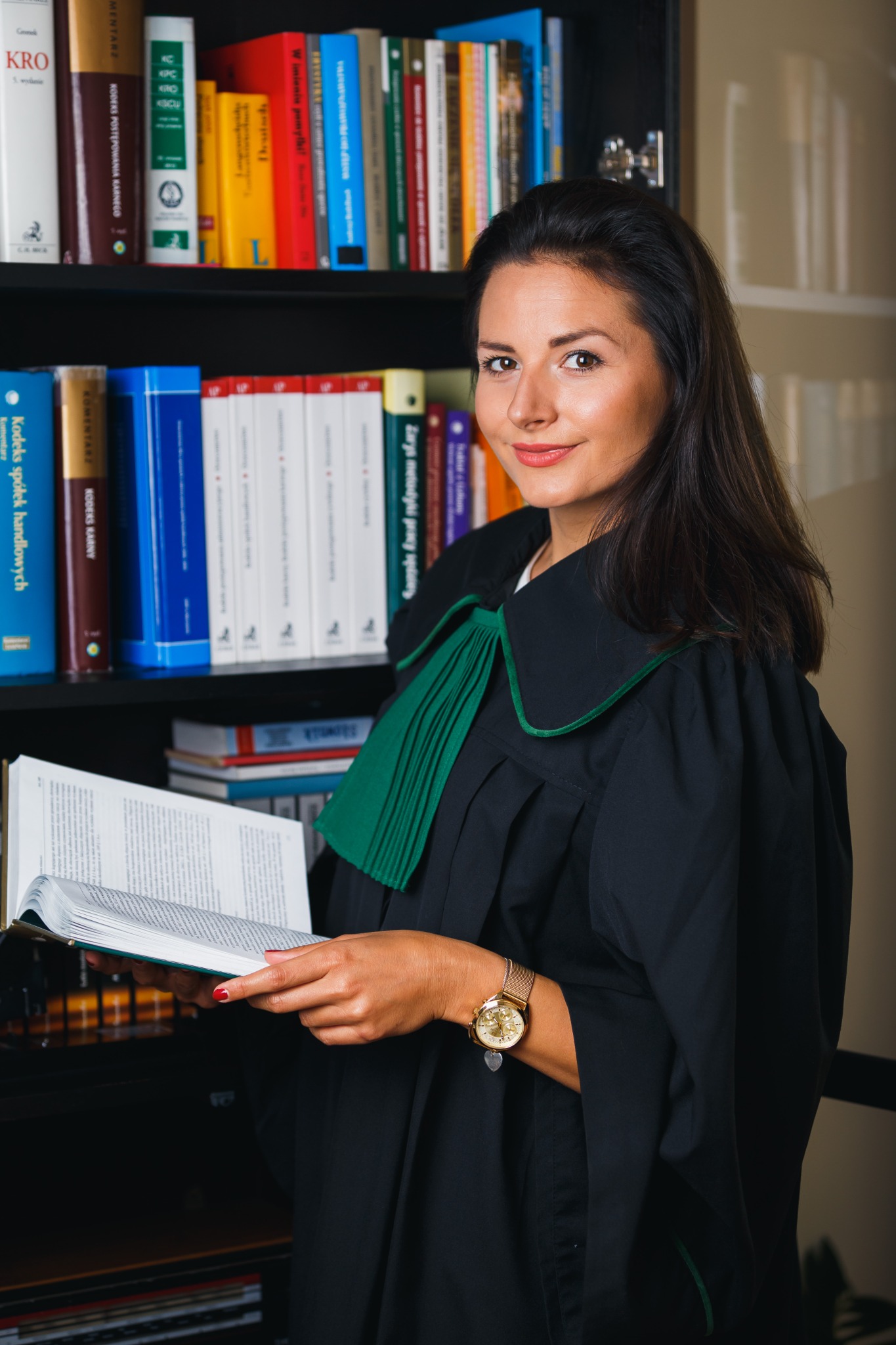 Adwokatka w todze, z uśmiechem, trzyma otwartą książkę na tle regału z książkami prawniczymi.