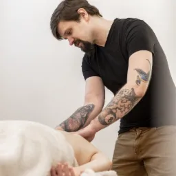 Tatuowany mężczyzna w czarnej koszulce wykonuje masaż pleców klientki leżącej na białym ręczniku.