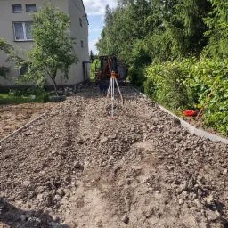 Przygotowanie podłoża z gruzem i kamieniami, widoczny niwelator, koparka w tle oraz obrzeża chodnikowe.