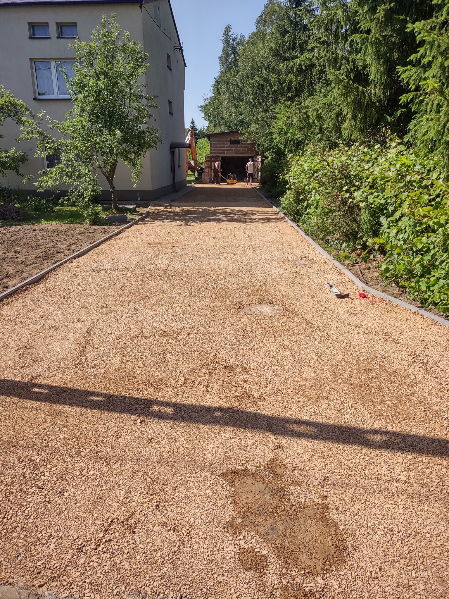 Nowo utwardzona droga z brązowego kruszywa, obrzeża z szarego betonu, w tle budynek z jasną elewacją, drzewa i pracujący robotnicy z koparką.