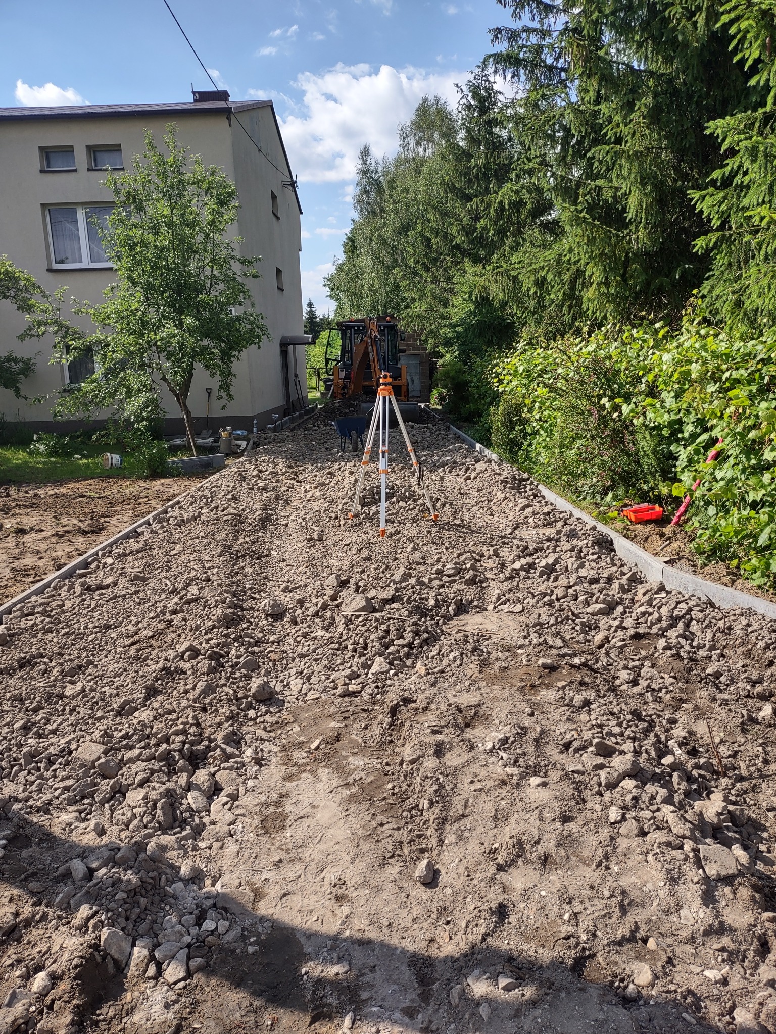 Przygotowanie podłoża z gruzem i kamieniami, widoczny niwelator, koparka w tle oraz obrzeża chodnikowe.
