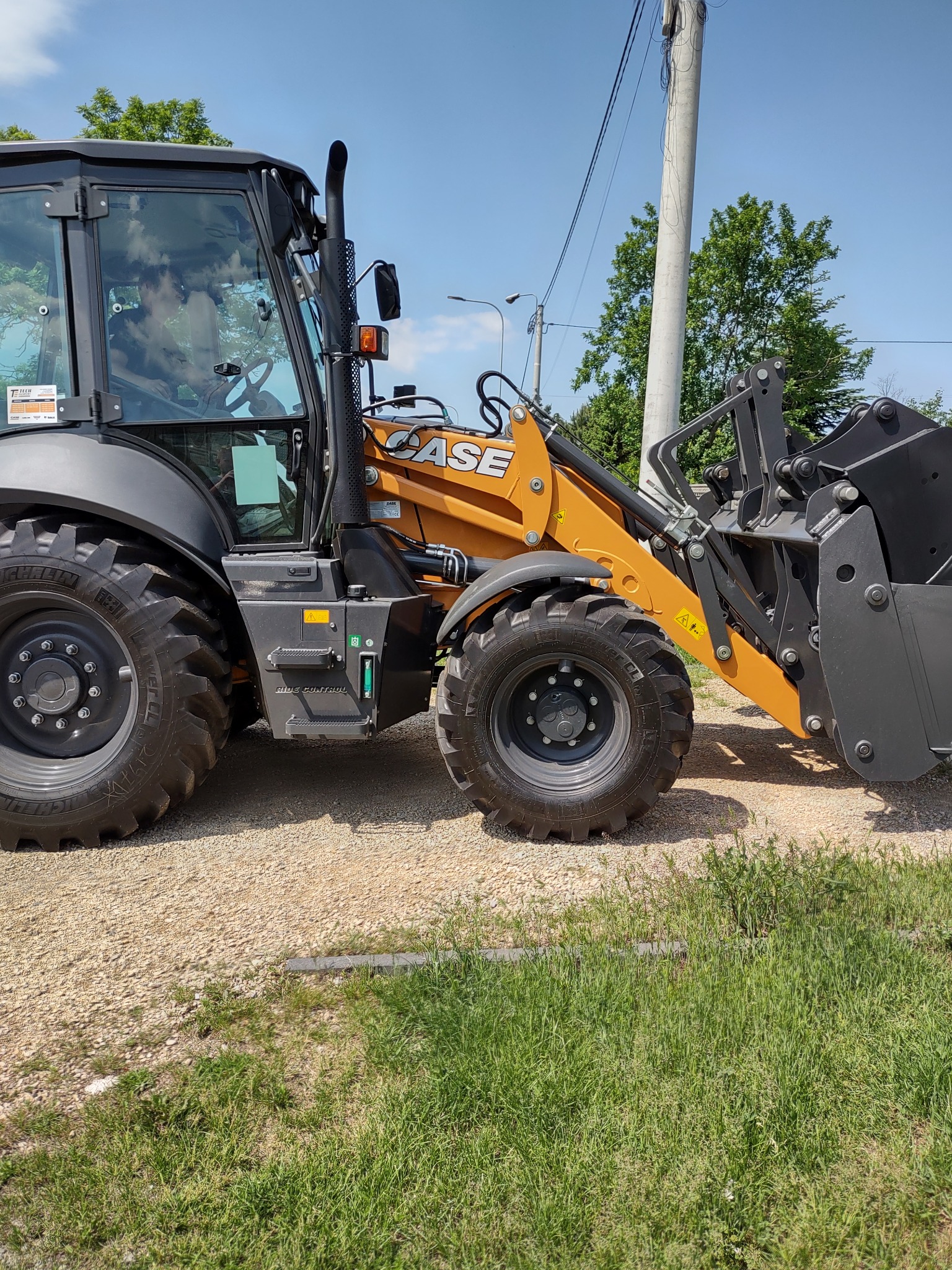 Pomarańczowo-szara koparko-ładowarka marki CASE na żwirowym podłożu, widok z boku, na tle słupa energetycznego i zieleni.