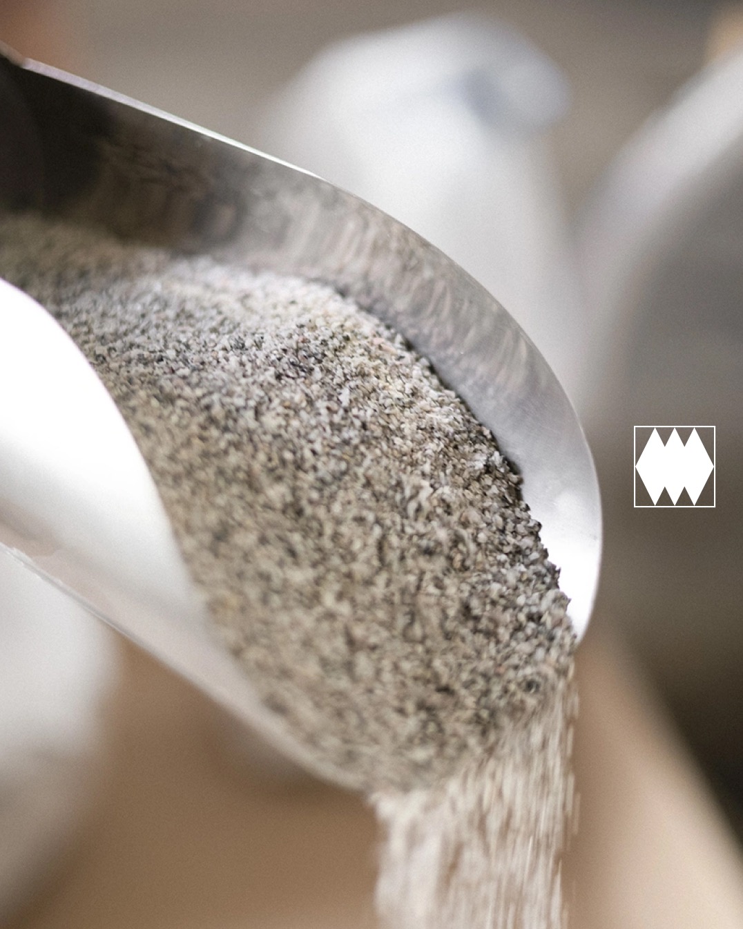 Metalowa łopatka nabiera szary, drobny materiał budowlany, który się z niej wysypuje. W tle widoczne pojemniki. W prawym rogu logo firmy.