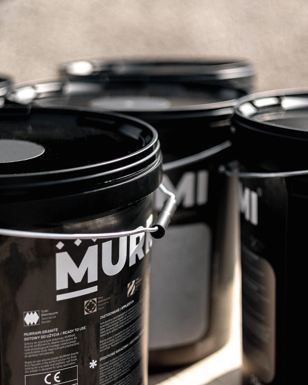 Czarne wiadra z tynkiem dekoracyjnym Murrami Granite, gotowym do użycia. Widoczne etykiety z informacjami o produkcie i zastosowaniu. Ujęcie z bliska, w tle kolejne wiadra.