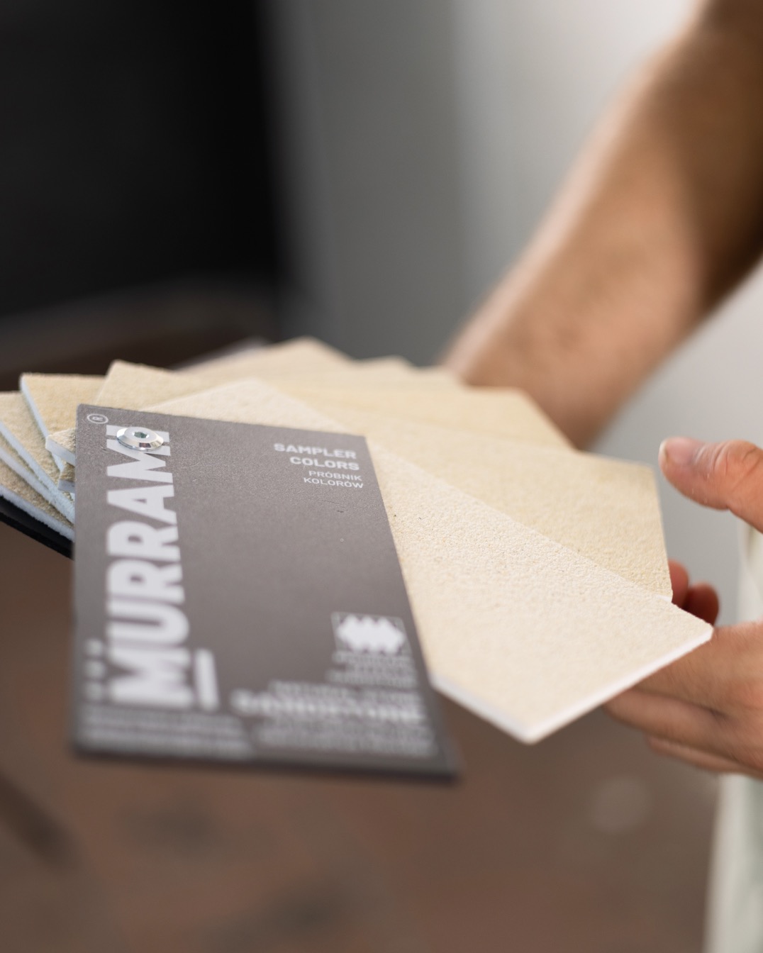 Próbniki kolorów i faktur Murram w dłoni, prezentacja palety beżowych odcieni. Karta z logo i napisem 'Sampler Colors'.