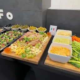 Stół cateringowy z przekąskami: mini kanapki z wołowiną i rukolą, deska serów i wędlin z winogronami, trzy rodzaje hummusu oraz pokrojone warzywa (ogórek, marchew) na drewnianych deskach, czarne...