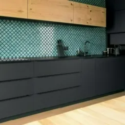 Nowoczesna kuchnia z ciemnoszarymi, matowymi frontami, blatem i turkusową mozaiką na ścianie, uzupełniona drewnianą półką wiszącą i jasną, drewnianą podłogą.