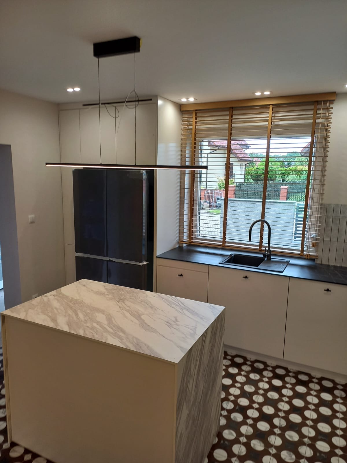 Nowoczesna kuchnia z wyspą o marmurowym blacie, minimalistycznym oświetleniem i drewnianymi żaluzjami w oknie. Uwagę zwraca wzorzysta podłoga i czarny zlew.