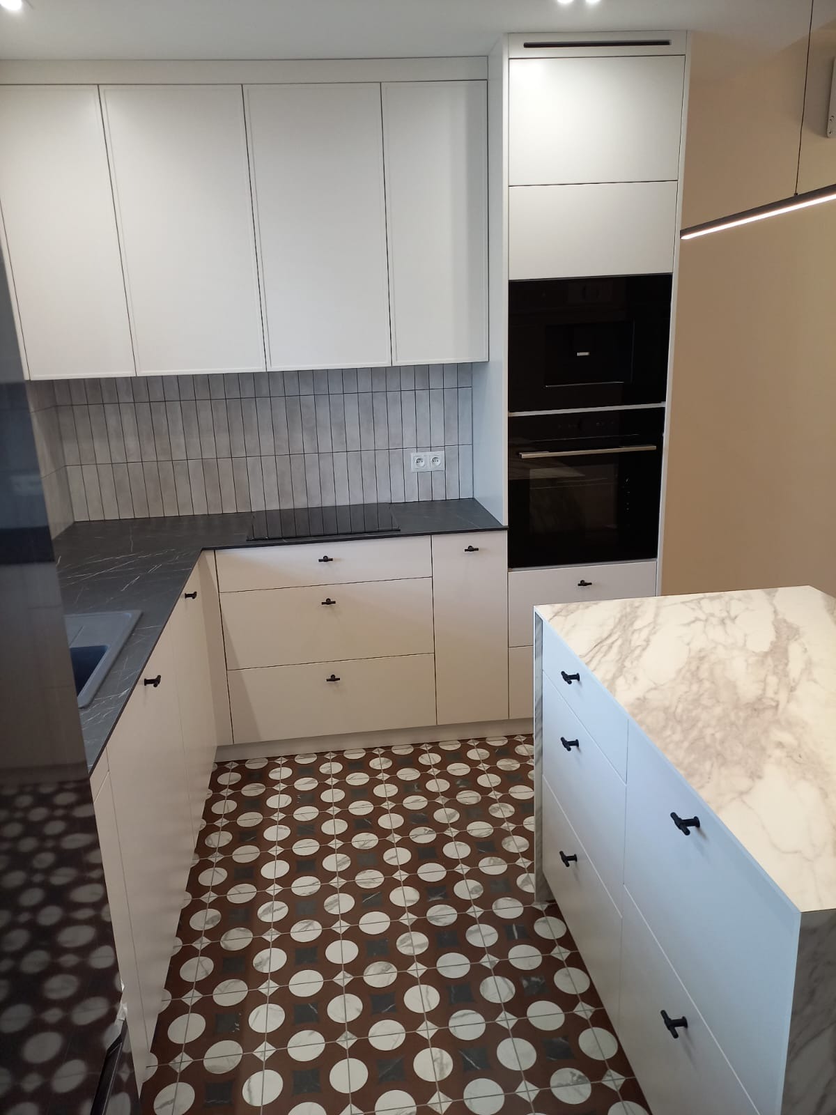 Nowoczesna, jasna kuchnia z wyspą i marmurowym blatem. Białe szafki z czarnymi uchwytami, szary blat i wzorzysta podłoga. Minimalistyczny design.