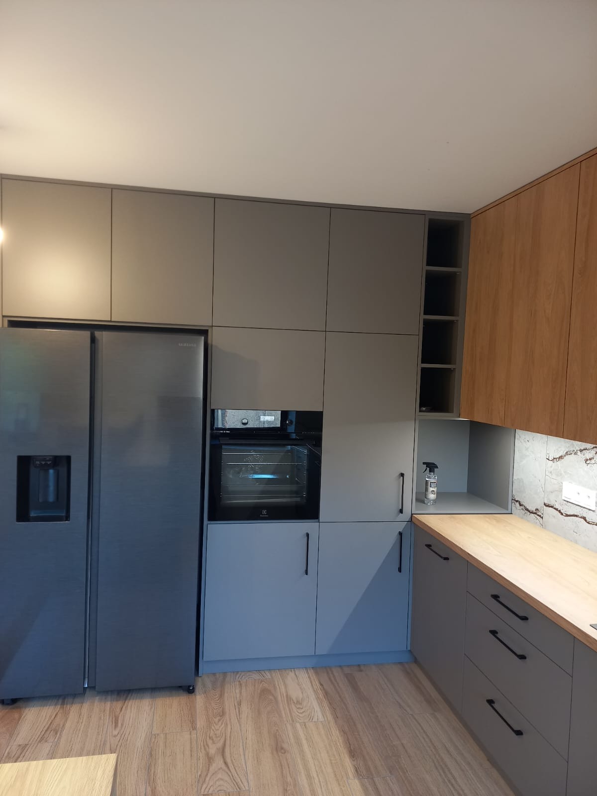 Nowoczesna kuchnia z szarymi szafkami, drewnianym blatem i lodówką side-by-side. Widoczny piekarnik w zabudowie i drewniane wykończenie górnych szafek.