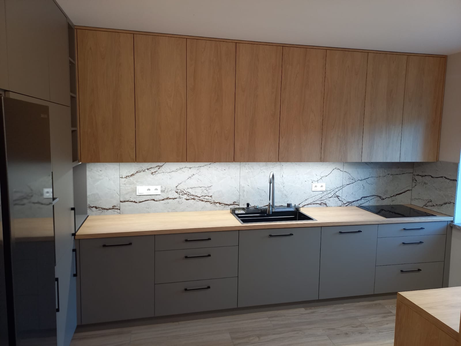 Nowoczesna kuchnia z szarymi szafkami dolnymi i drewnianymi górnymi, blatem w kolorze jasnego drewna i marmurowym backsplash'em. Minimalistyczne uchwyty.