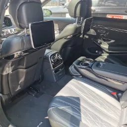 Wnętrze luksusowego samochodu Mercedes S klasa AMG 63S z widocznymi skórzanymi fotelami z pikowaniem, ekranami w zagłówkach, konsolą środkową z uchwytami na napoje i dywanikami z logo AMG.