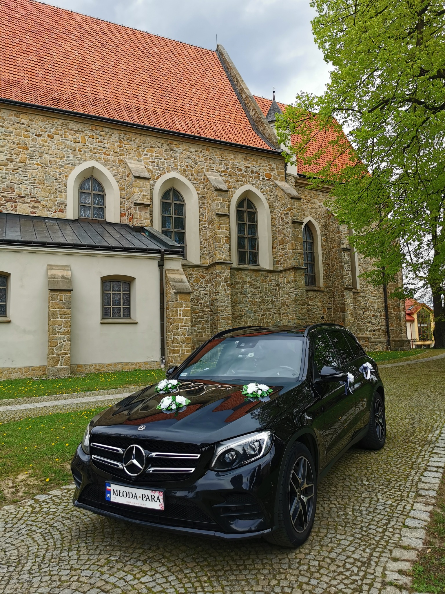 Czarny Mercedes GLC udekorowany białymi kwiatami i tablicą rejestracyjną 'Młoda Para' na tle kamiennego kościoła z czerwoną dachówką.