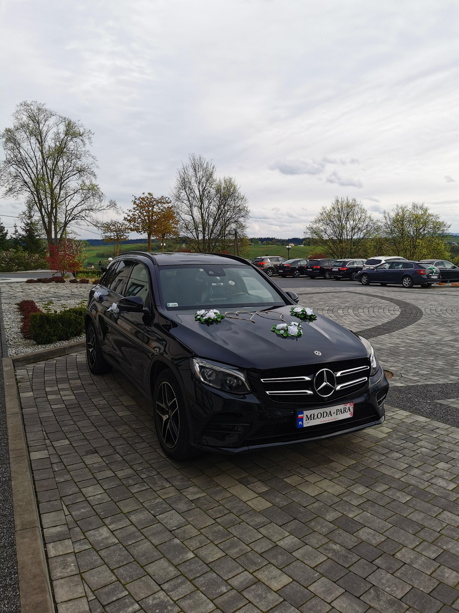 Czarny Mercedes GLC 220d 4 matic, udekorowany kwiatami i tablicą 'Młoda Para', zaparkowany na brukowanym parkingu w Rzeszowie, z widokiem na zalesione wzgórza w tle.