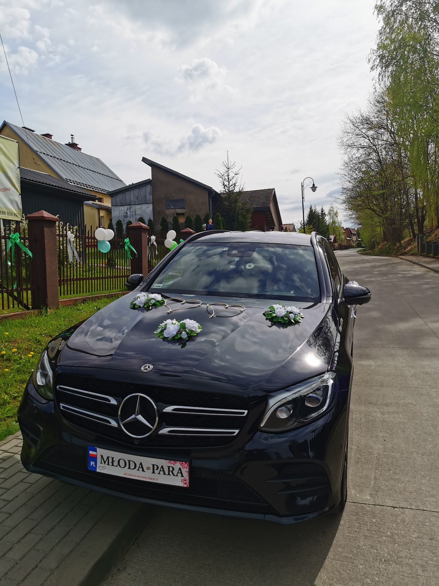 Czarny Mercedes GLC 220d 4 matic udekorowany kwiatami i ozdobami ślubnymi, zaparkowany na ulicy przed domem z ogrodzeniem ozdobionym balonami i wstążkami.