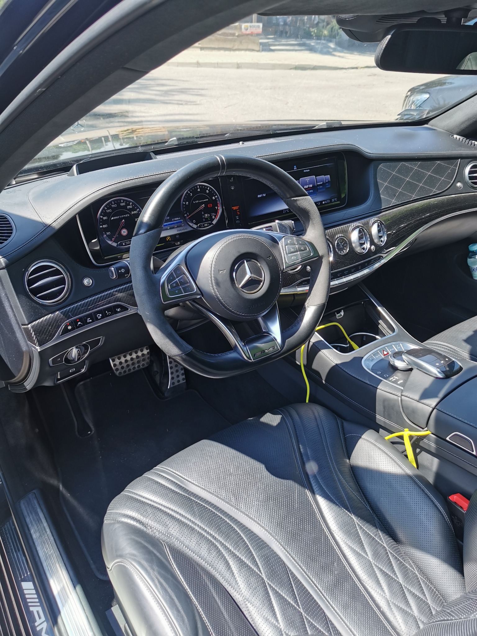 Wnętrze luksusowego samochodu Mercedes S klasa AMG 63S z czarną skórzaną tapicerką, widok na deskę rozdzielczą, kierownicę z logo AMG i elementy wykończenia z włókna węglowego, z żółtym kablem...