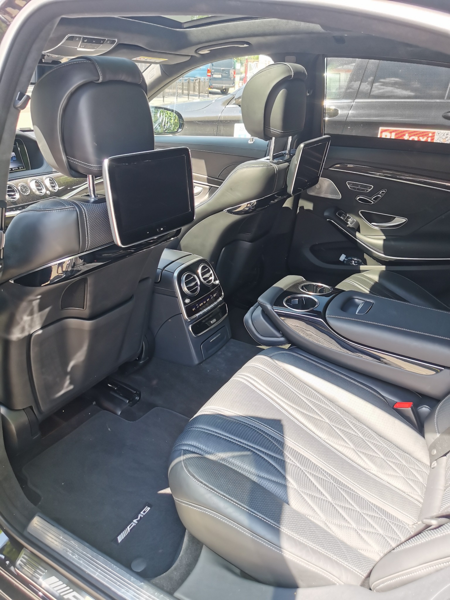 Wnętrze luksusowego samochodu Mercedes S klasa AMG 63S z widocznymi skórzanymi fotelami z pikowaniem, ekranami w zagłówkach, konsolą środkową z uchwytami na napoje i dywanikami z logo AMG.