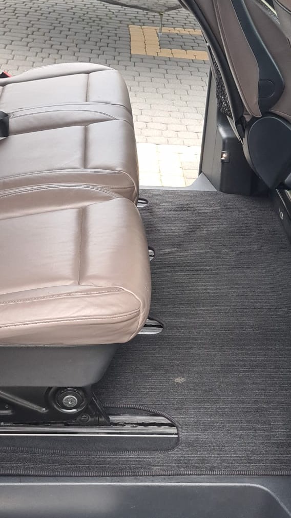 Wnętrze pojazdu z beżową skórzaną kanapą i szarym dywanikiem podłogowym, widok z boku na szyny mocujące siedzenia.