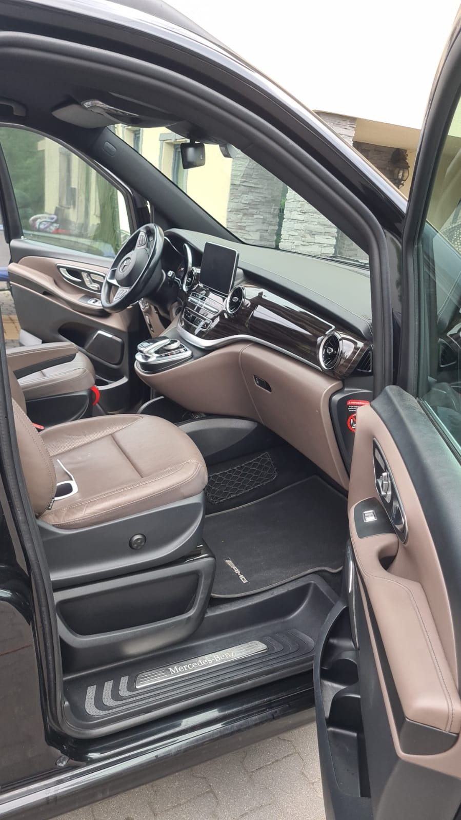 Wnętrze czarnego luksusowego vana Mercedes-Benz z brązową skórzaną tapicerką, widoczne fotele, deska rozdzielcza z drewnianymi akcentami i otwarte drzwi.