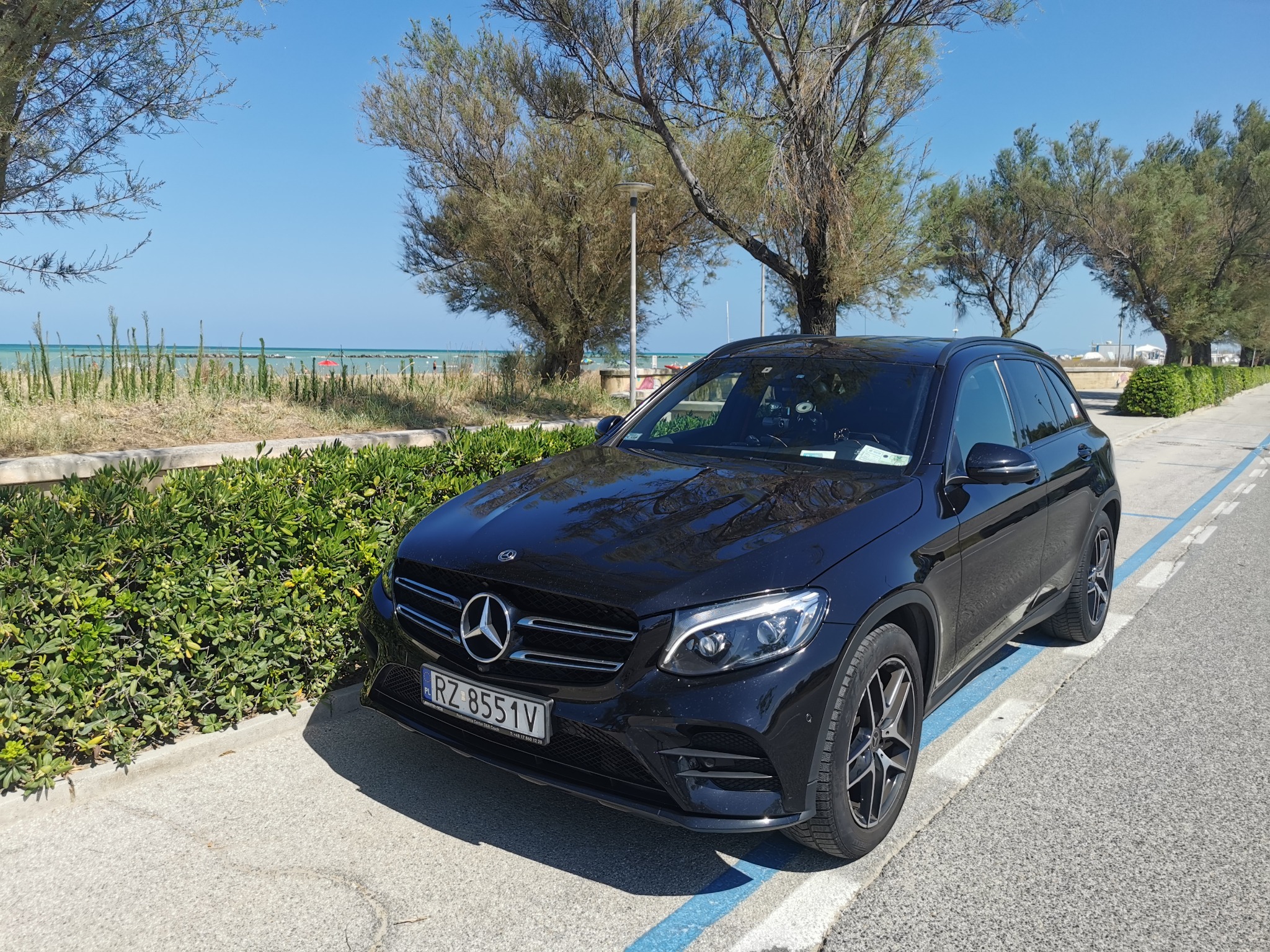 Czarny Mercedes GLC 220d 4matic zaparkowany na parkingu z niebieską linią, w tle krzewy i drzewa, w oddali widoczne morze i plaża.