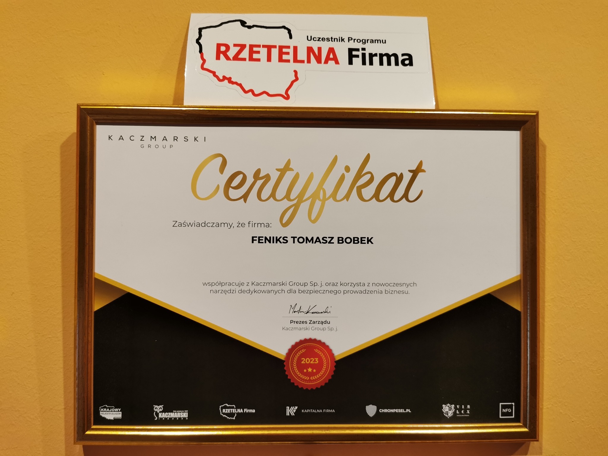 Oprawiony w złotą ramę certyfikat firmy FENIKS TOMASZ BOBEK, potwierdzający współpracę z Kaczmarski Group Sp. j. i korzystanie z nowoczesnych narzędzi biznesowych, obok naklejka 'Rzetelna Firma'.