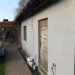 Świeżo otynkowana ściana budynku z brązową stolarką okienną i drewnianymi drzwiami o geometrycznym wzorze, widoczne rynny, zawór wody i narzędzia malarskie obok ścieżki z kostki.