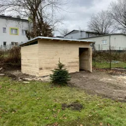Surowa drewniana konstrukcja małego budynku gospodarczego z otwartymi drzwiami, na trawiastym terenie z małą choinką przed budynkiem, w tle budynki mieszkalne i pochmurne niebo.